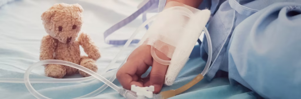Child's hand covered in medical bandages and tubes with a small brown teddy bear next to them.