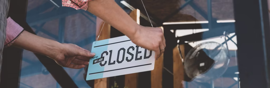 A person places a 'We Are Closed' sign on a clear glass door. Business Interruption featured image